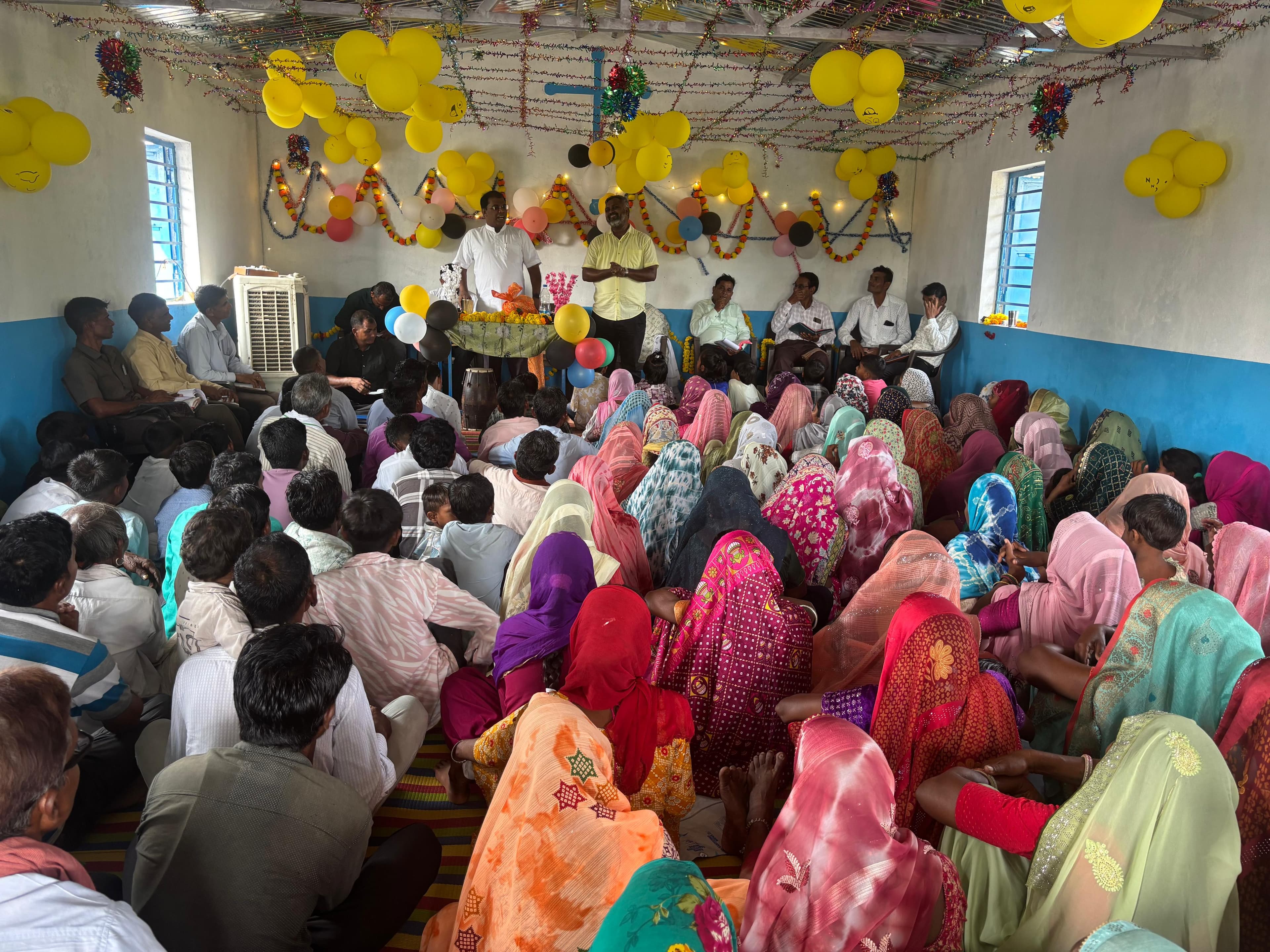 A congregation seated on the floor of a decorated hall during a gathering.